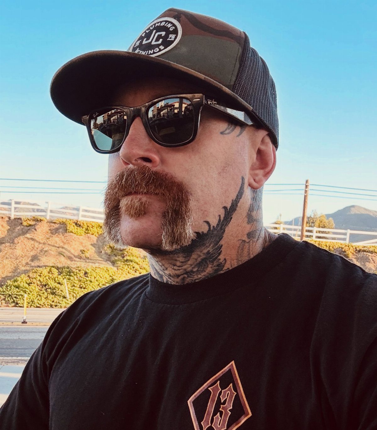 Man with a brown horseshoe mustache wearing sunglasses and a camouflage cap.