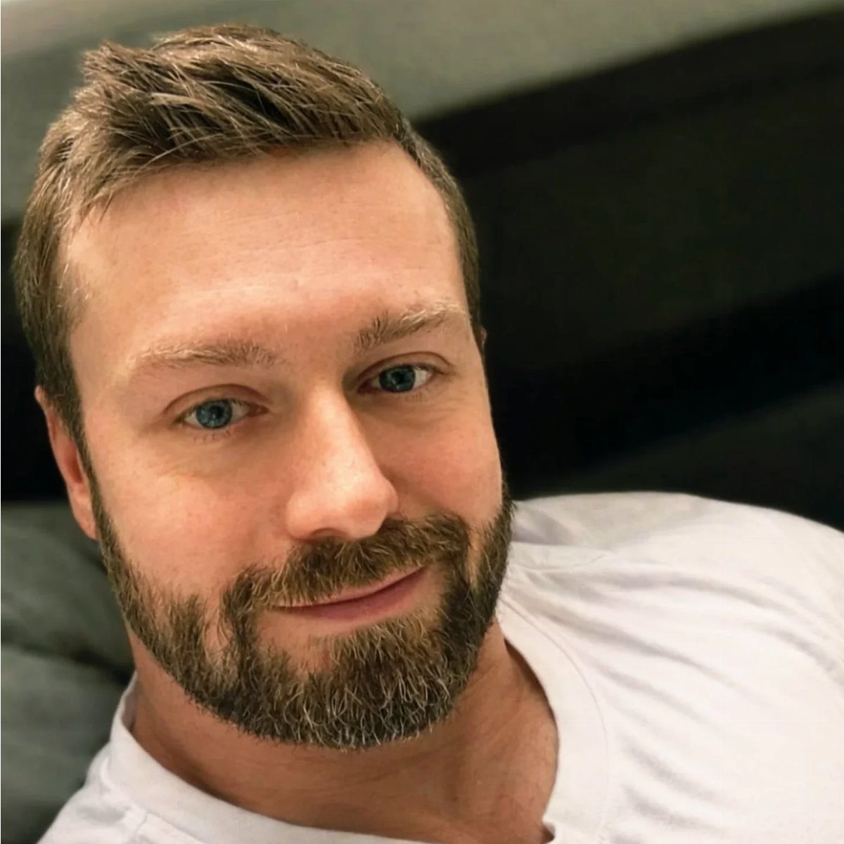 Man with a full, neatly trimmed brown beard and mustache wearing a white shirt.