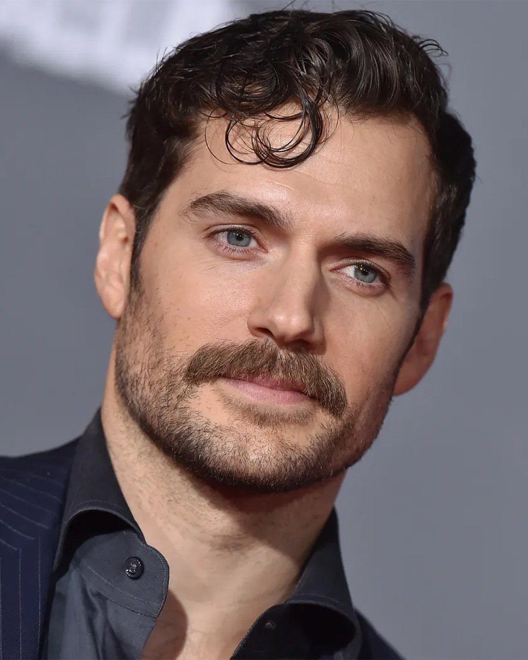 Man with a thick brown mustache and short stubble beard wearing a dark suit.