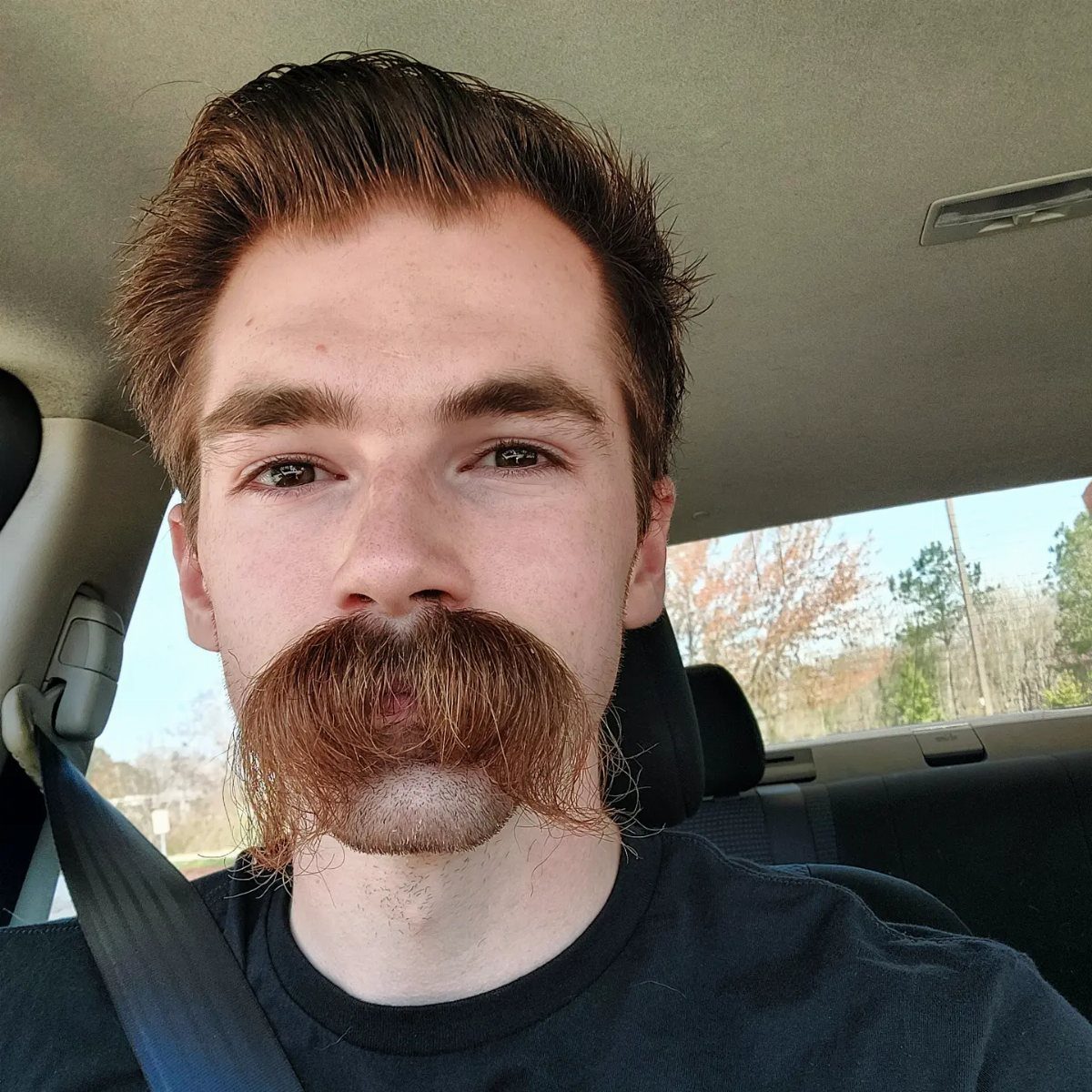 Man with a large, bushy walrus mustache sitting inside a car.