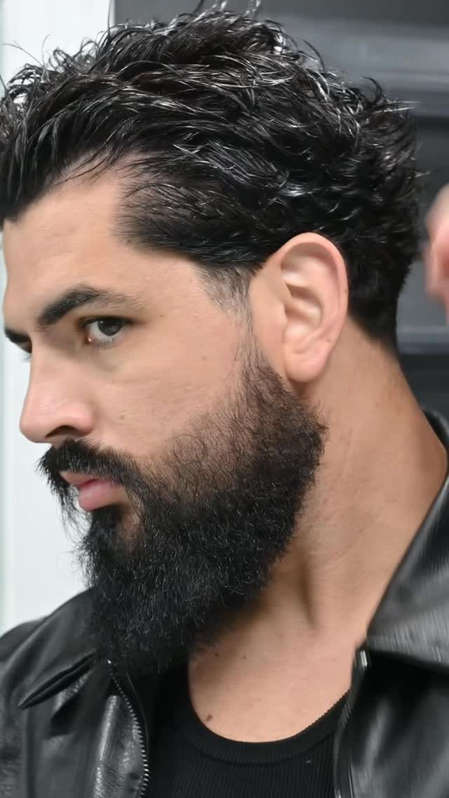 Man with a thick dark full beard and wavy hair wearing a leather jacket.