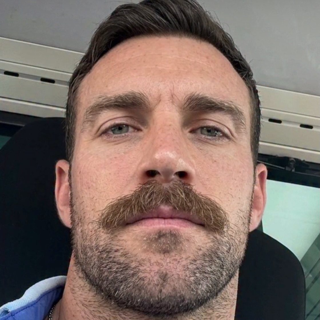 Man with a thick brown chevron mustache and short facial stubble.
