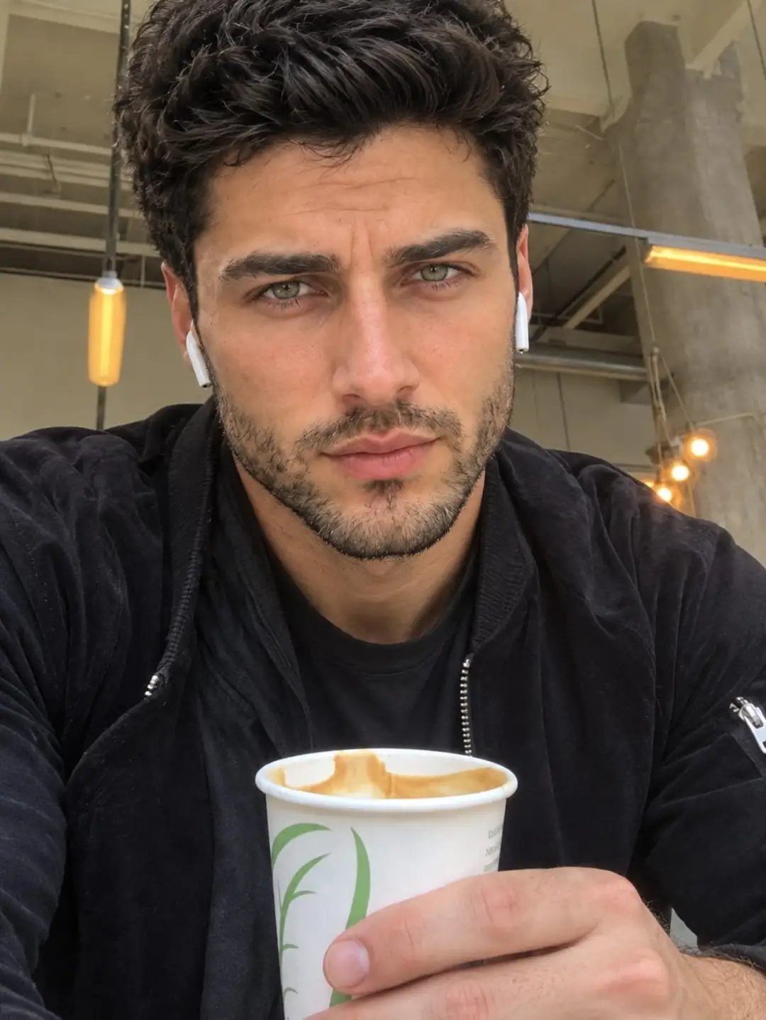 Man with short dark stubble beard holding a white coffee cup indoors.