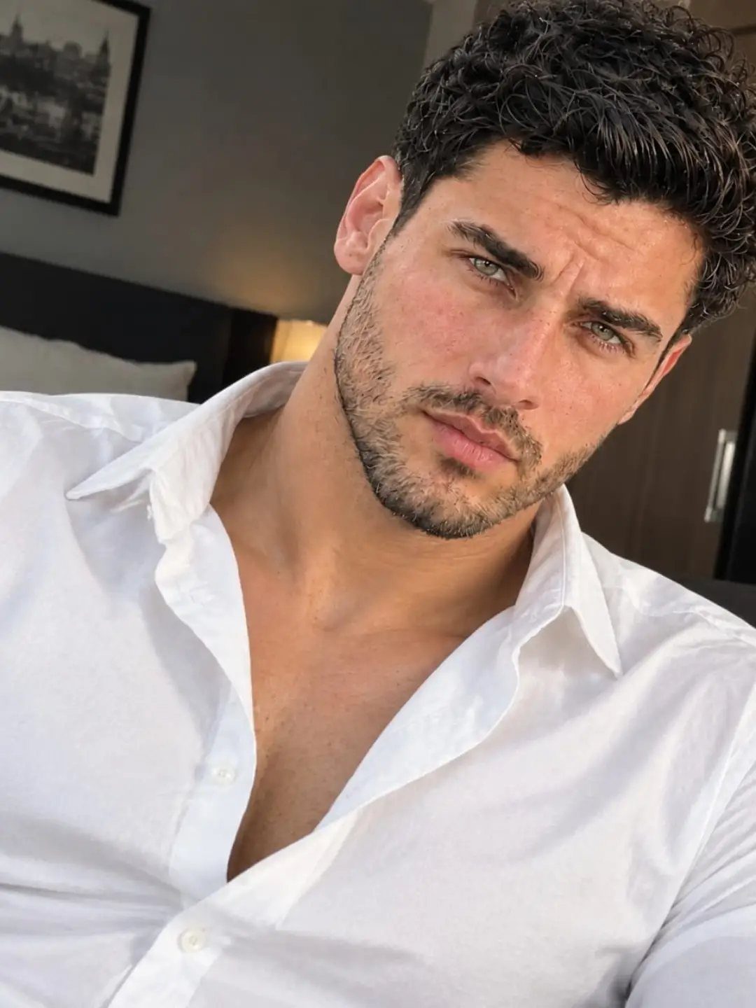 Man with dark curly hair and medium stubble wearing a white shirt.