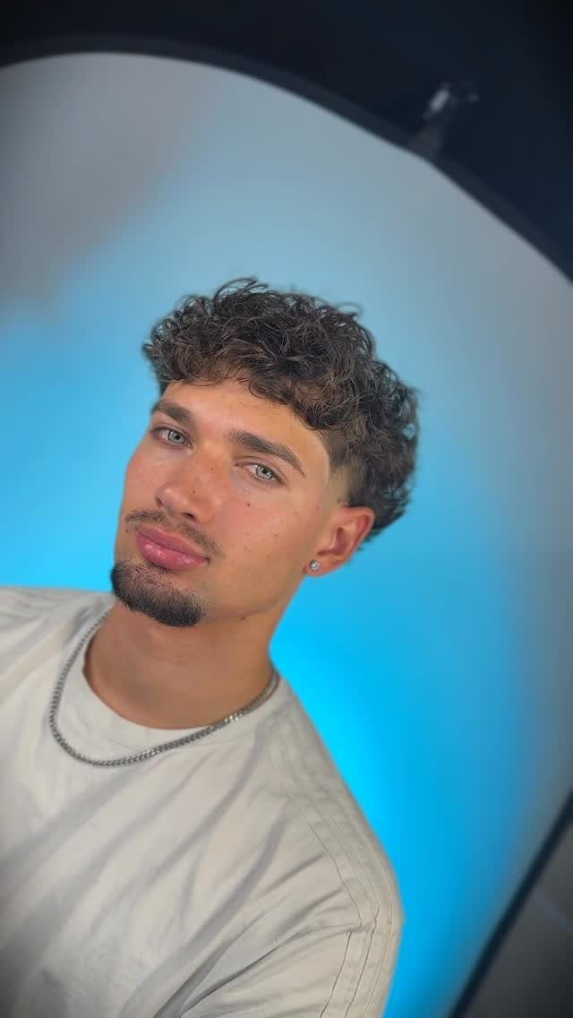 Young man with curly fringe haircut and tapered sides posing against blue light.