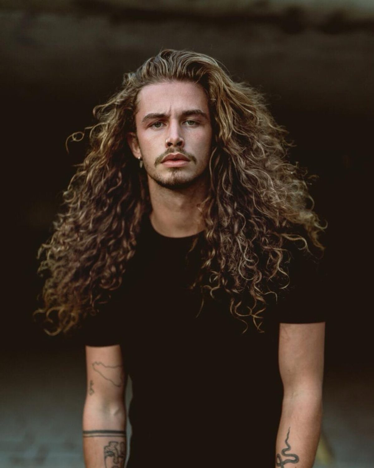 Man with long voluminous curly brown hair and a mustache wearing a black shirt.