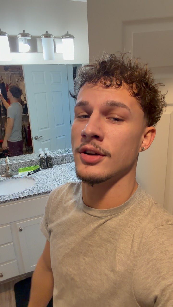 Young man with short curly brown hair and mustache posing in a bathroom