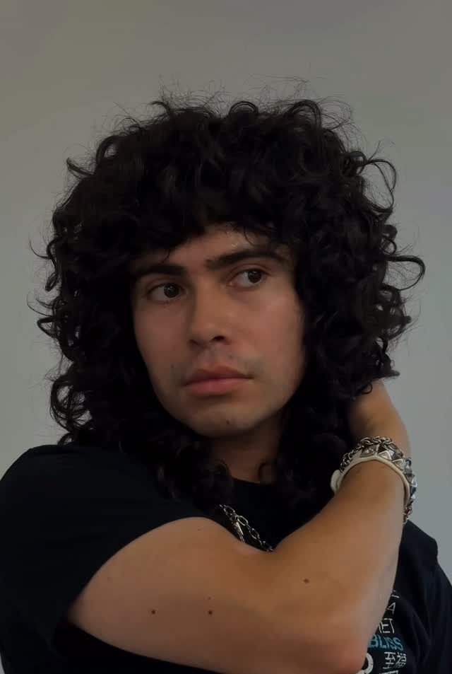 Man with voluminous shoulder-length dark curly hair posing against a grey wall.