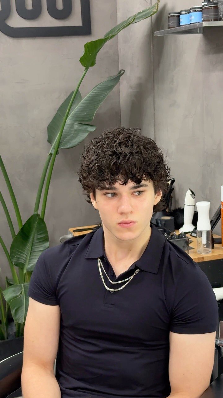 Young man with voluminous dark brown curly hair wearing a black polo shirt.