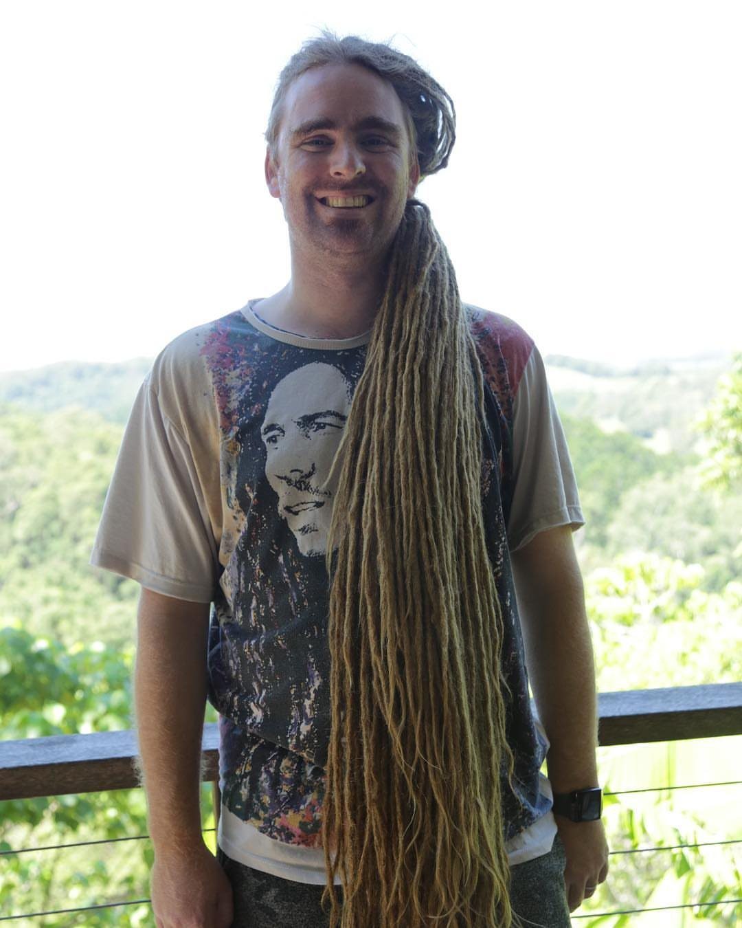 Man with very long blonde dreadlocks smiling on a balcony overlooking green hills