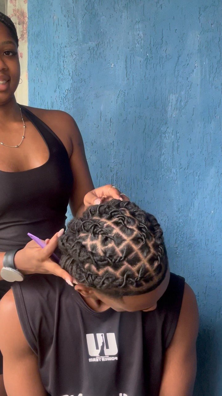 Man receiving a hairstyle of thick criss-crossed dreadlock twists against a blue wall