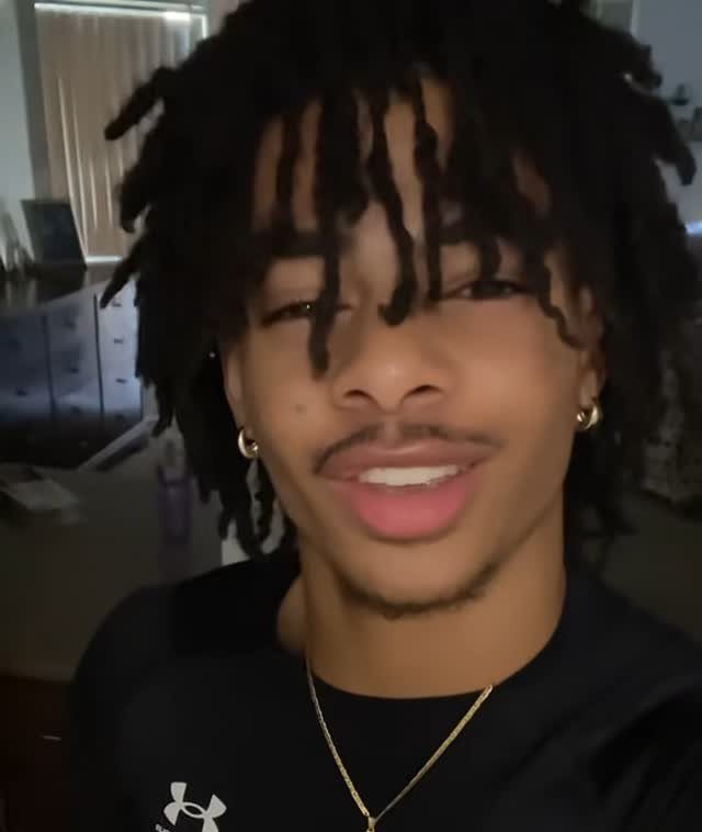 Young man with black dreadlocks, gold earrings, and a black t-shirt posing indoors.
