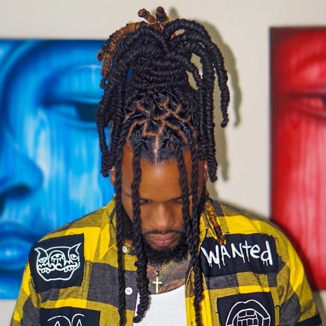 Man with long dark dreadlocks wearing a yellow plaid shirt against abstract art.