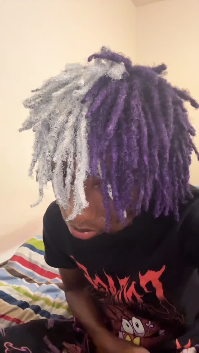 Man with split-color dreadlocks in silver and purple wearing a black graphic t-shirt.
