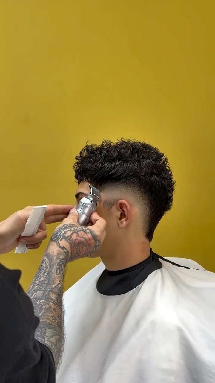Barber cutting a burst fade on a man with curly hair against yellow wall