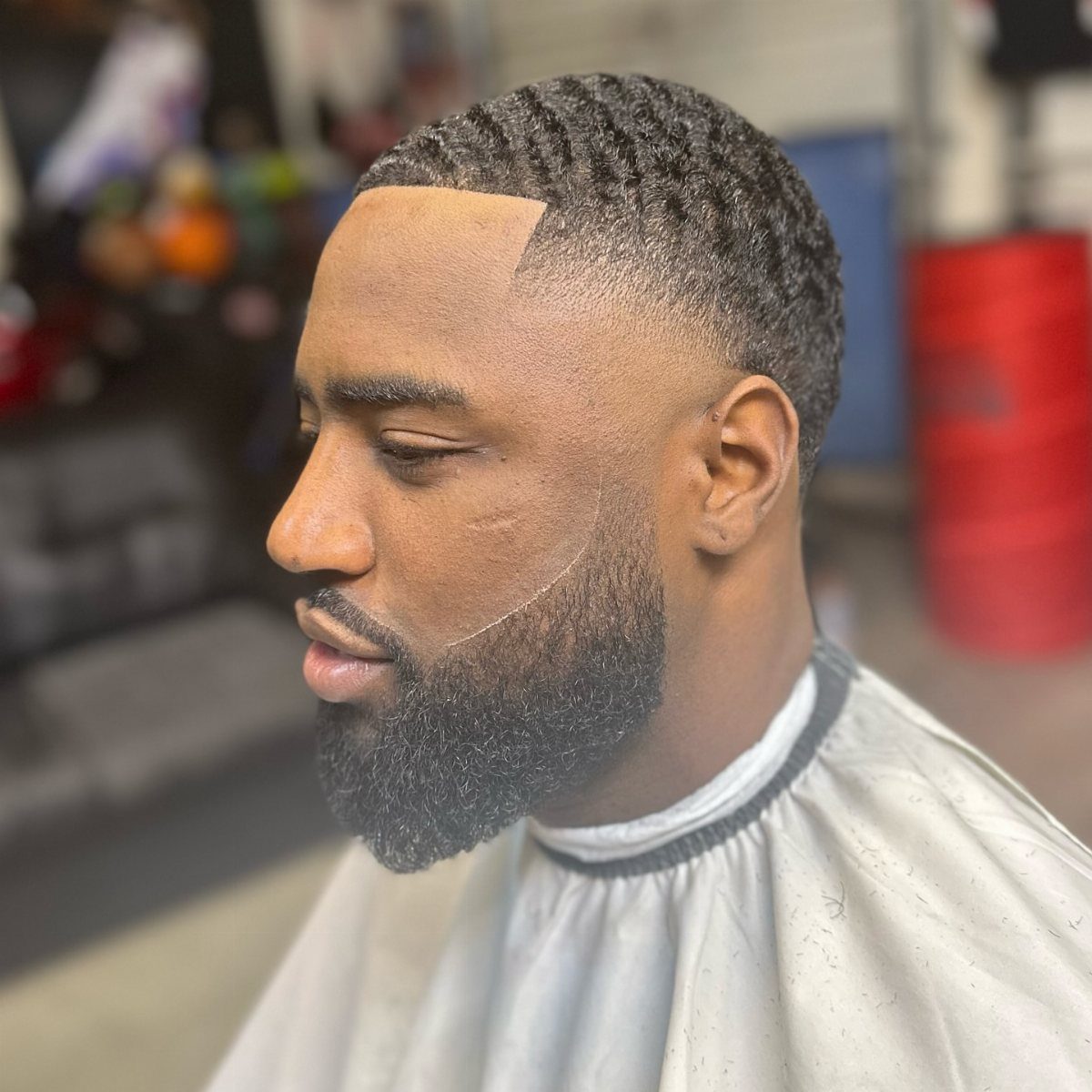 Man with a temple fade haircut and a groomed black beard wearing a cape.