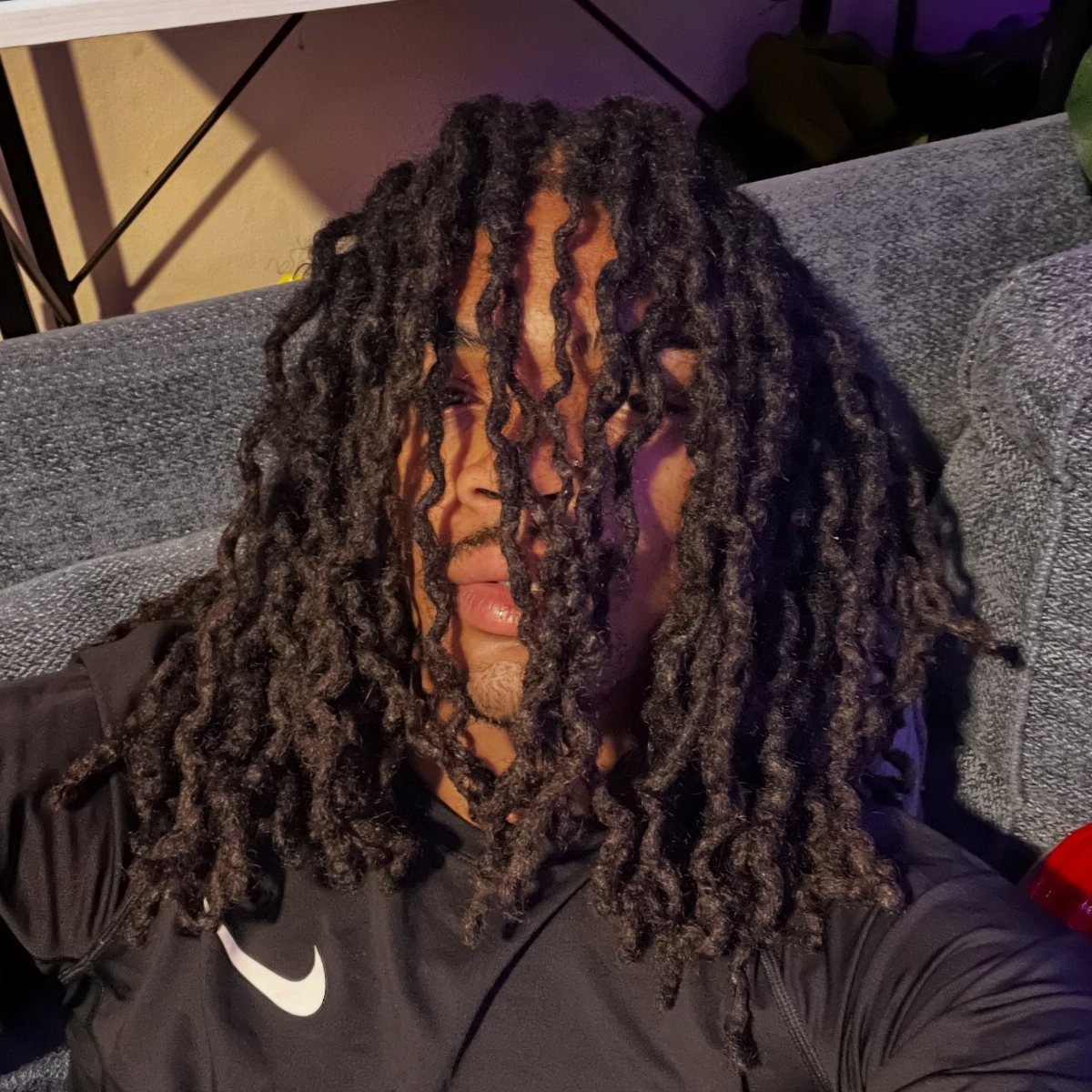 Man with long dark brown dreadlocks wearing a black Nike shirt on a sofa.