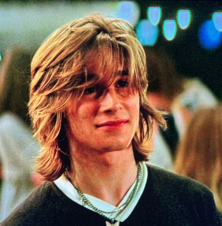 Young man with long blonde hair styled in a center part and layers.