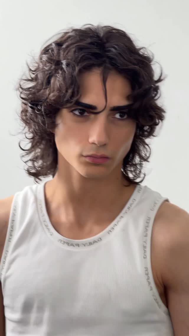 Young man with long, dark, wavy hair wearing a white ribbed tank top.