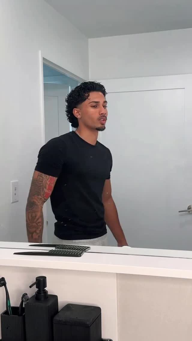 Man with dark curly hair slicked back reflecting in a bathroom mirror.