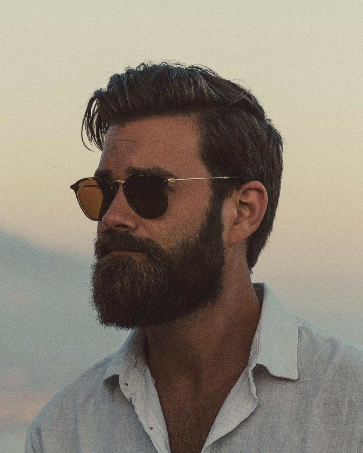 Man with a full brown beard and styled hair wearing sunglasses and white shirt.