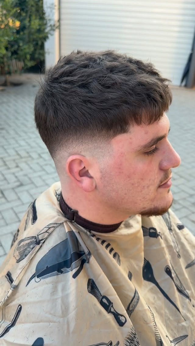 Man with a textured French crop haircut and skin fade wearing a barber cape.