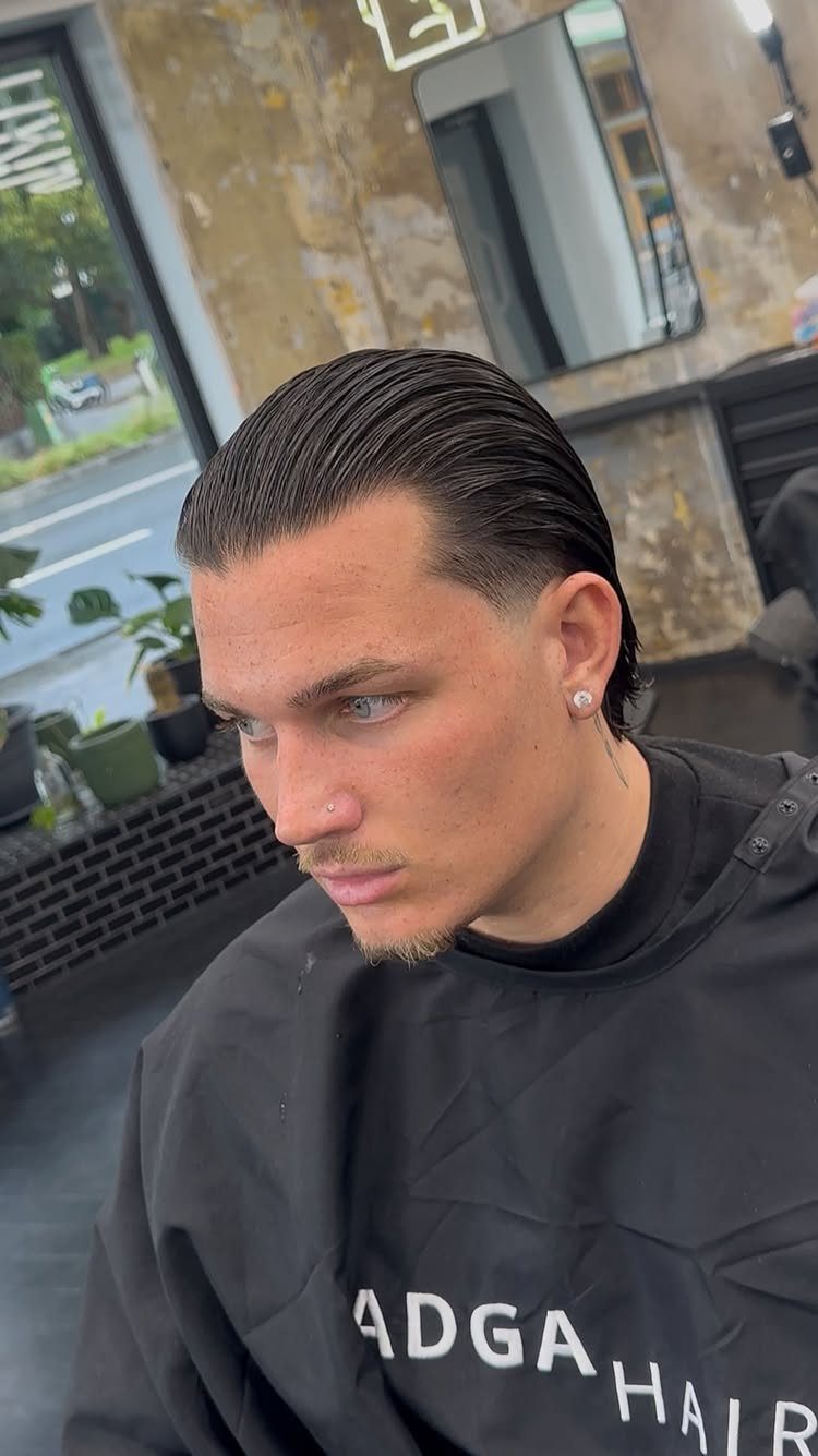 Man with a slicked-back undercut hairstyle wearing a black barber cape in a salon.