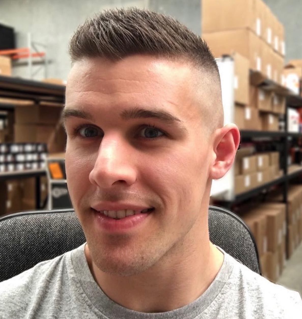 Man with a high fade haircut smiling in a warehouse with cardboard boxes.