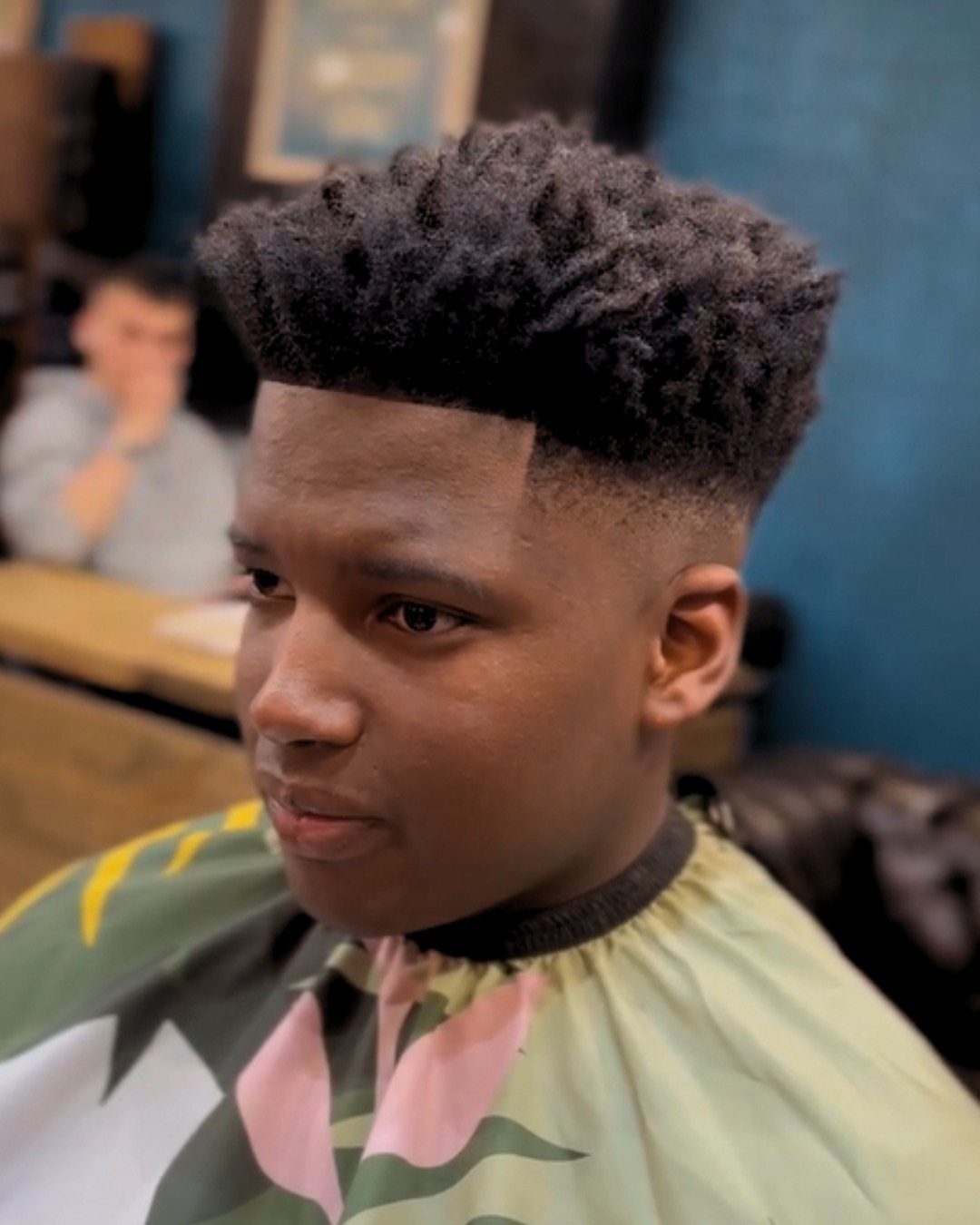 Young man with a high fade afro and textured hair wearing a barber cape.