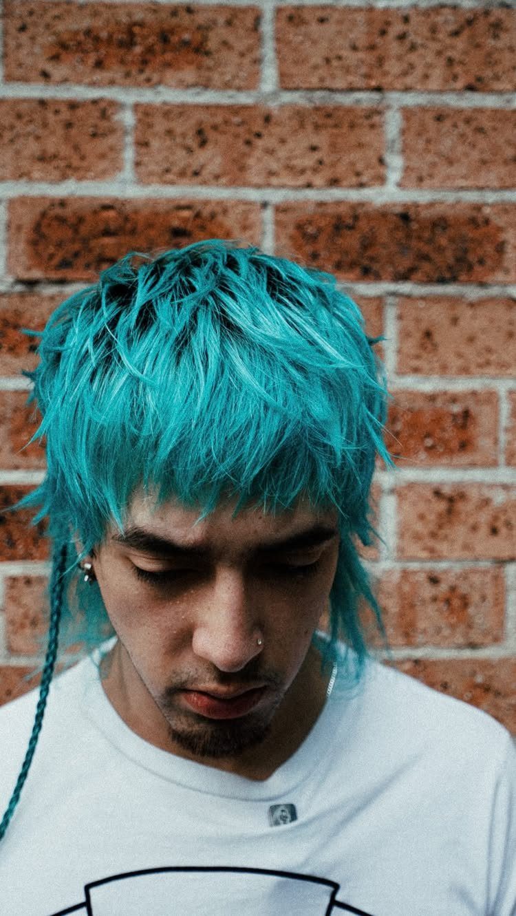 Man with bright turquoise choppy layered hair and bangs against a brick wall