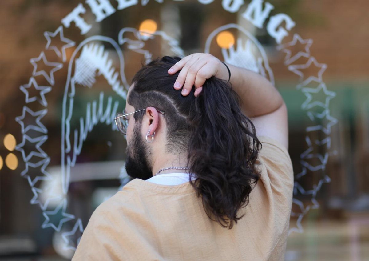 Man with long dark wavy hair and shaved sides wearing a tan shirt.