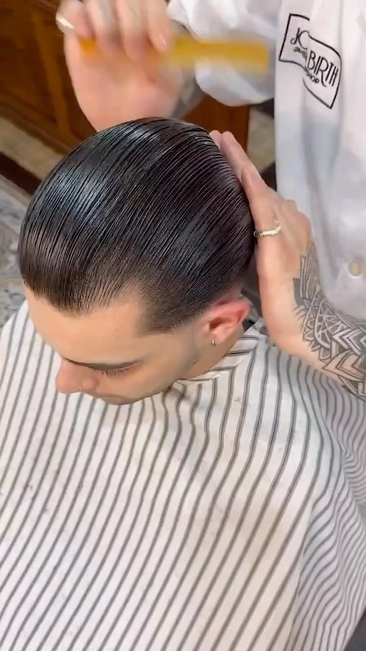Man with dark straight hair being slicked back by a barber in a salon