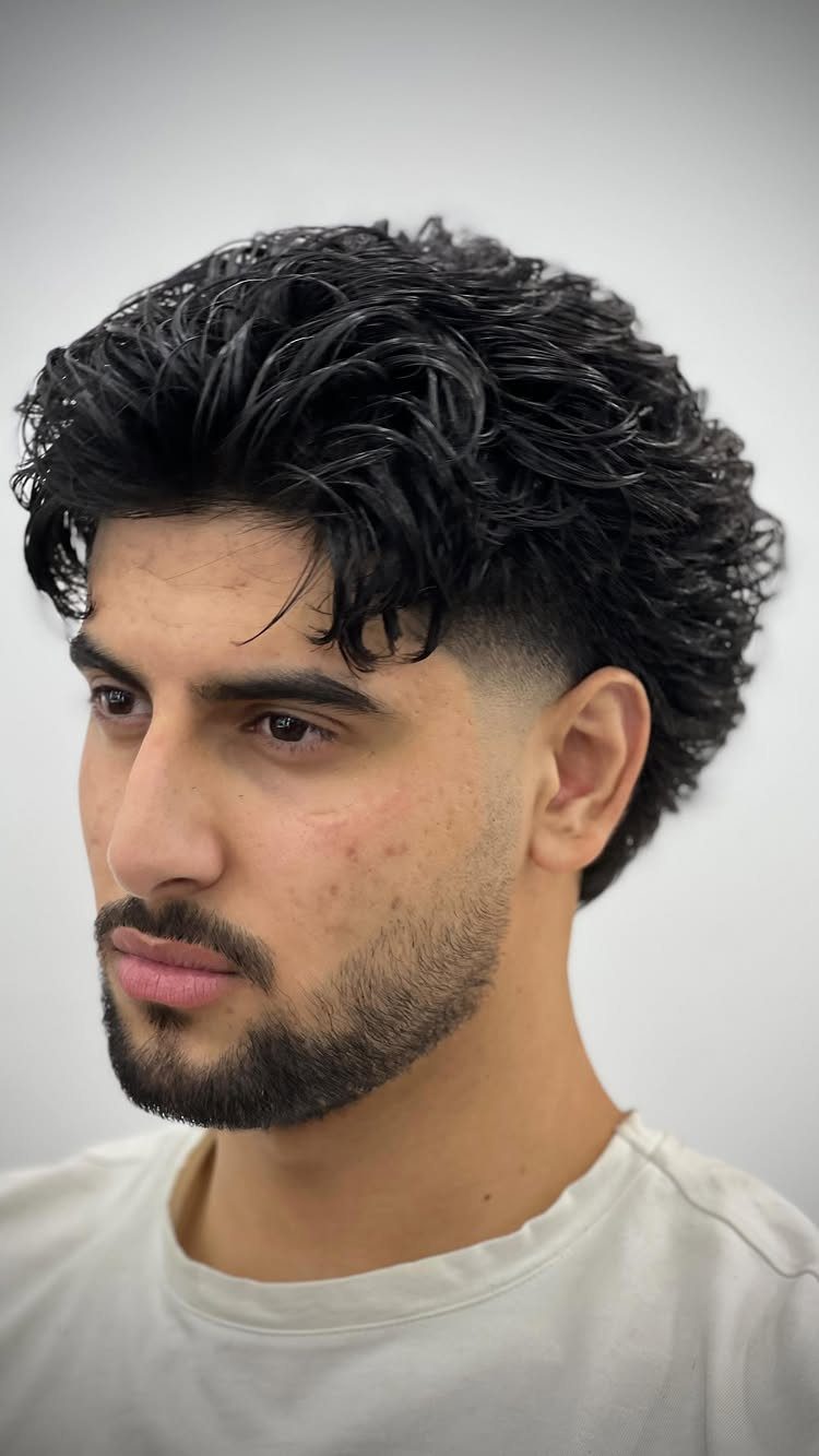 Man with a low fade mullet haircut and curly dark hair and beard.