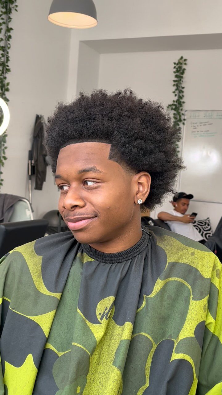 Young man with a rounded black afro and clean hairline wearing a barber cape.