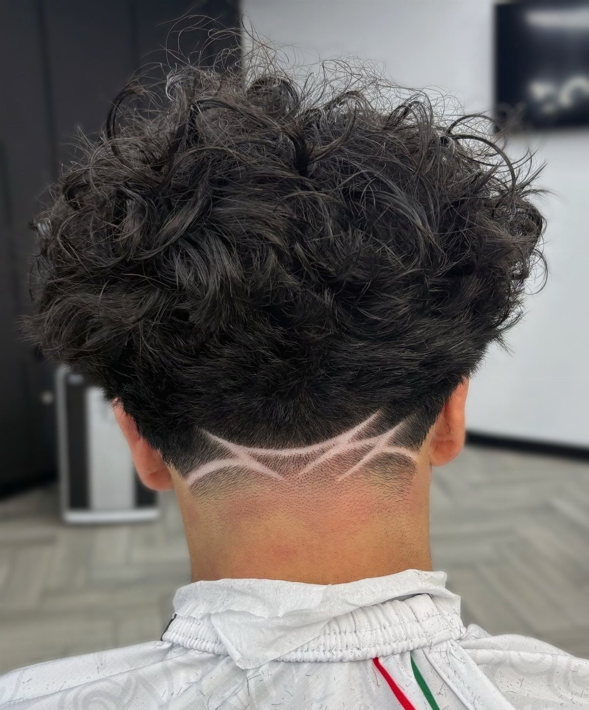 Curly dark brown hair with a low taper fade and shaved curved lines.