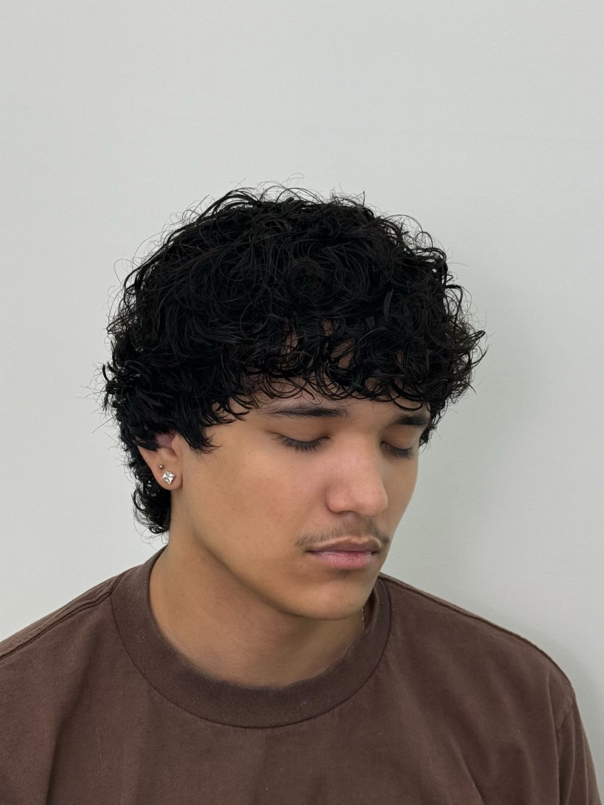 Young man with curly black hair and a brown t-shirt against white wall