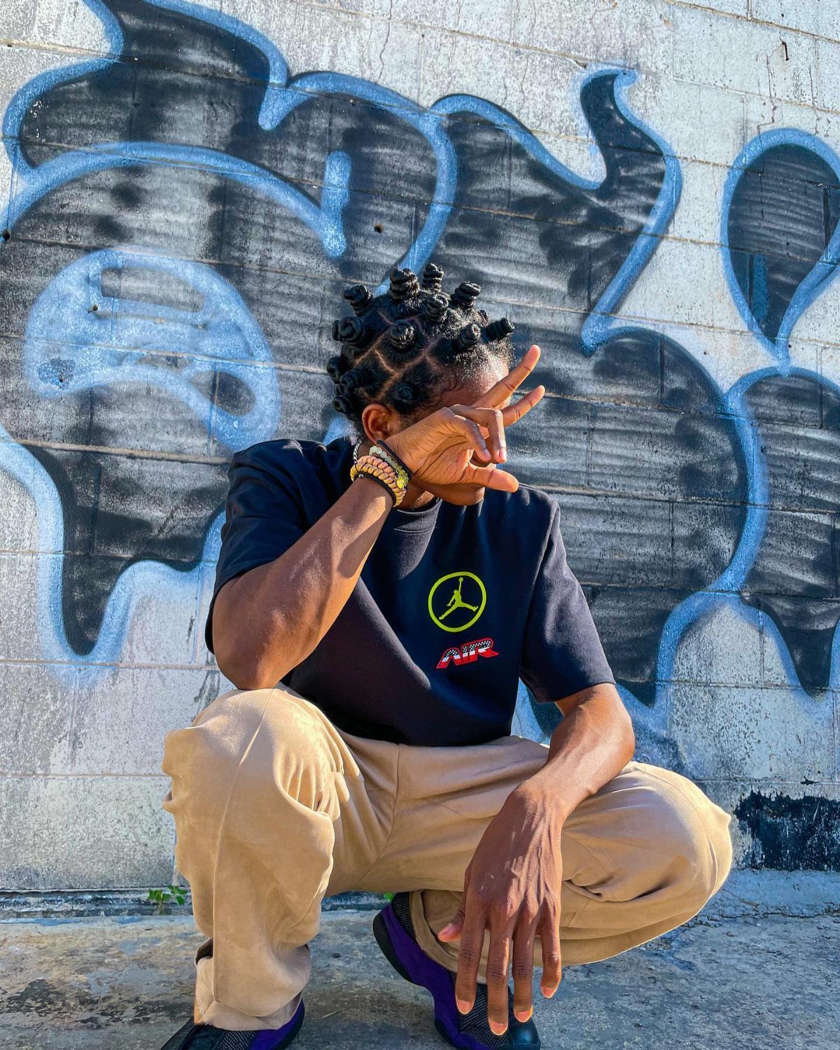 Man with medium length twisted hair crouching before a blue graffiti wall.