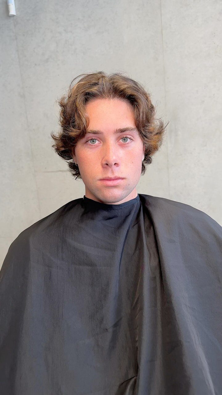 Young man with medium length textured wavy brown hair wearing a black barber cape.