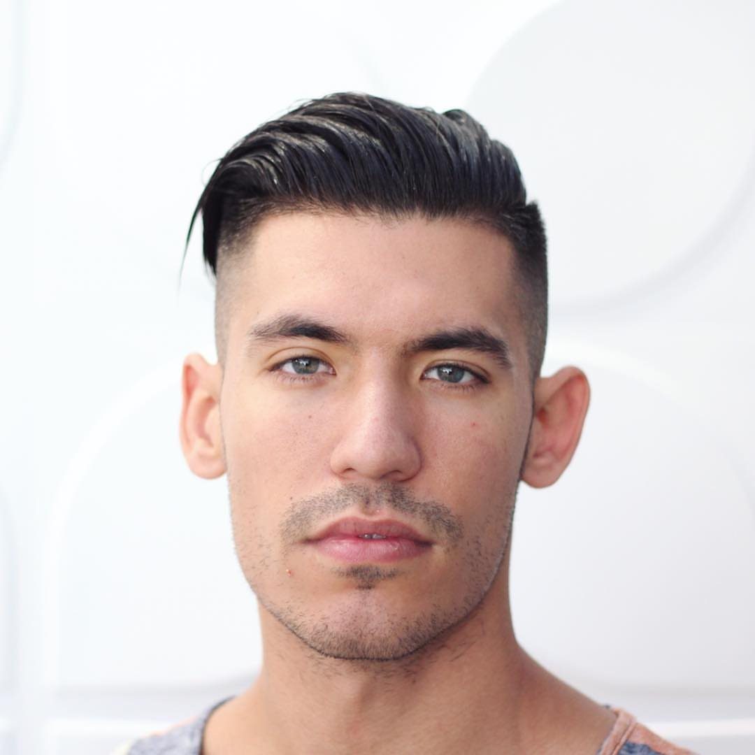 Man with a dark undercut hairstyle and slicked-back top against a white background.