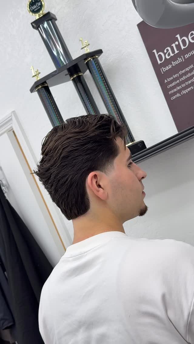 Man with dark straight hair slicked back in a tapered cut at a barbershop