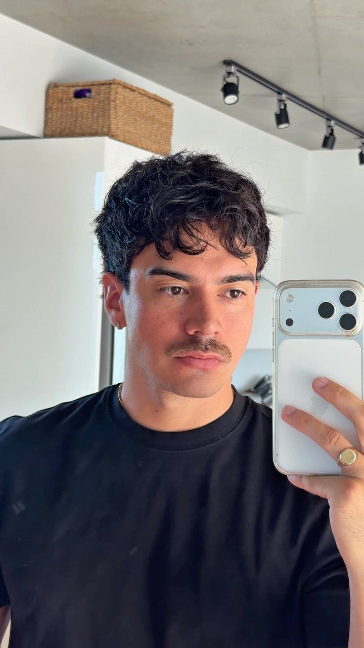 Man with short dark curly hair and a mustache taking a mirror selfie.