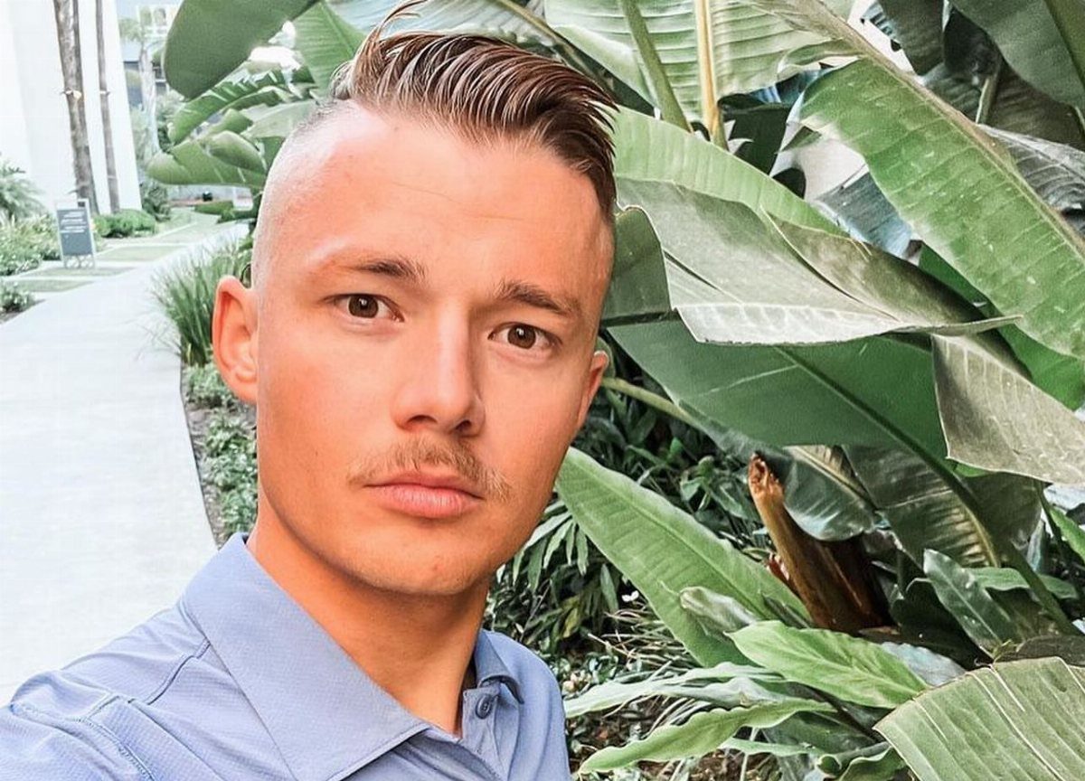 Man with an undercut side part hairstyle posing against large green tropical leaves.
