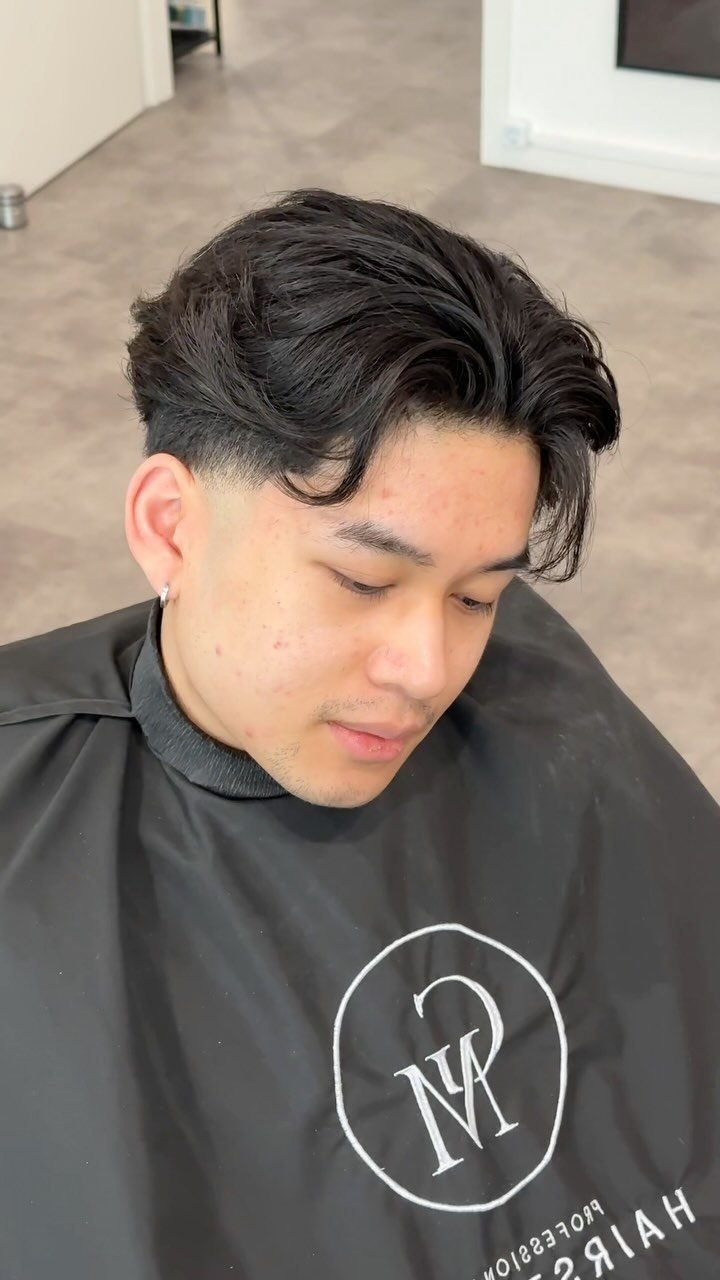 Man with a side-swept comb over fade haircut wearing a black barber cape.