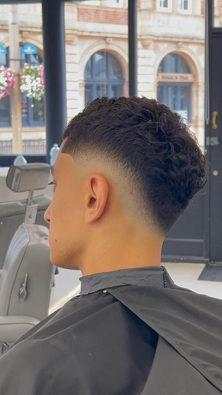 Man with a curly drop fade haircut wearing a black barber cape.