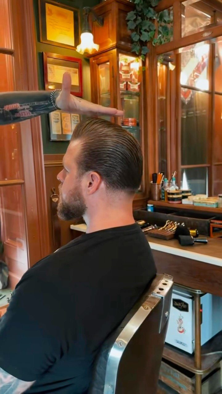 Man with slicked-back hair and beard in a classic wooden barbershop setting.