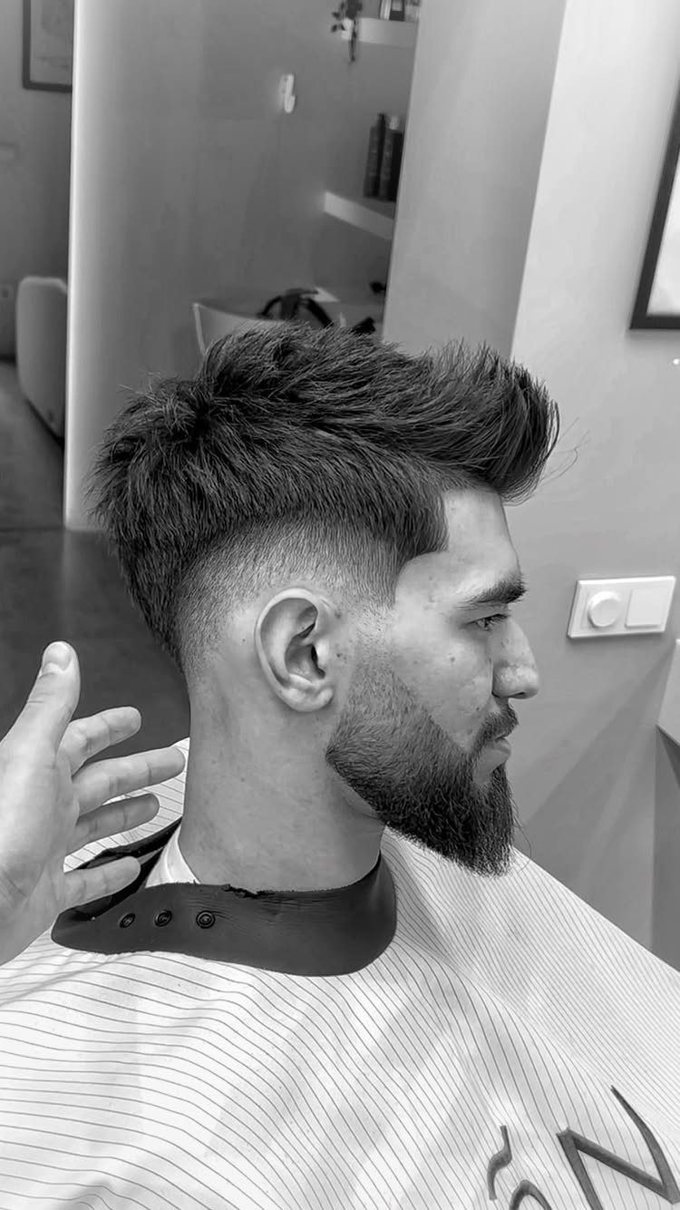 Man with a quiff haircut and beard wearing a barber cape in profile.
