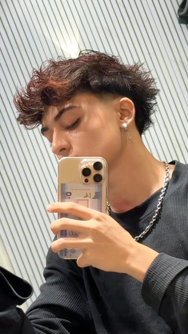 Young man with wavy reddish-brown hair and shaved sides taking a mirror selfie.