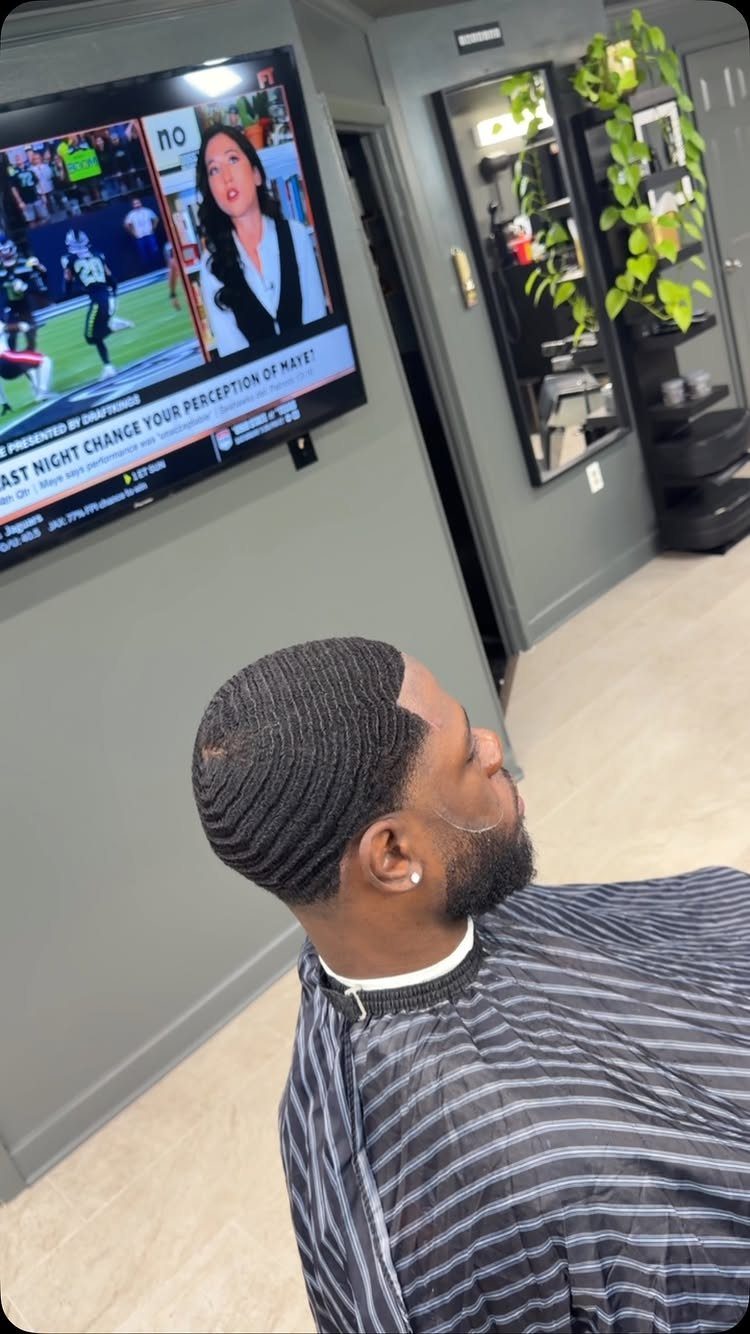 Man with 360 waves and a beard wearing a barber cape in a shop.