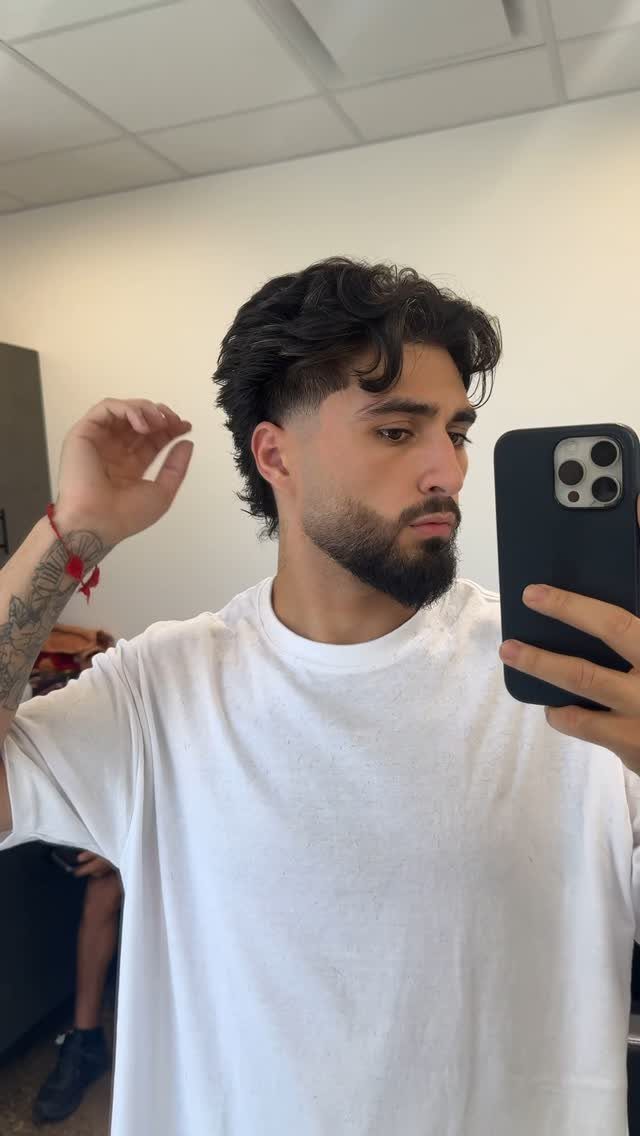 Man with dark wavy hair and a beard taking a mirror selfie.