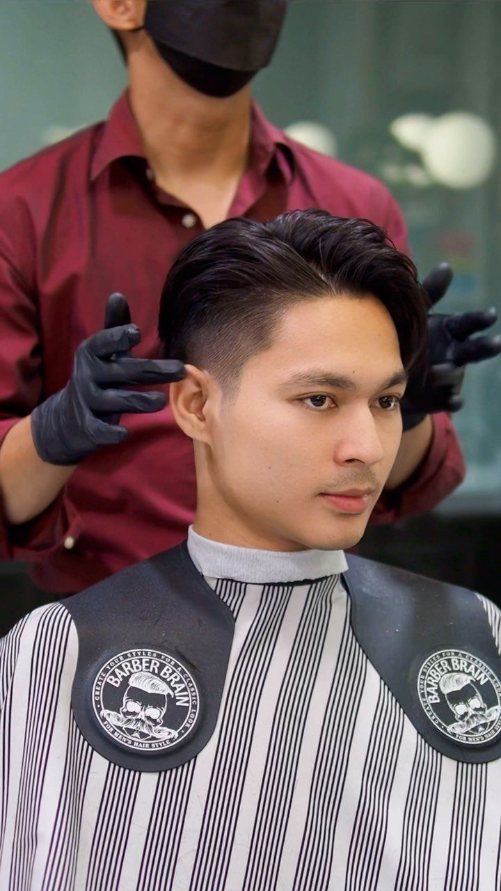 Man with a slicked-back undercut hairstyle being groomed by a barber in a shop.